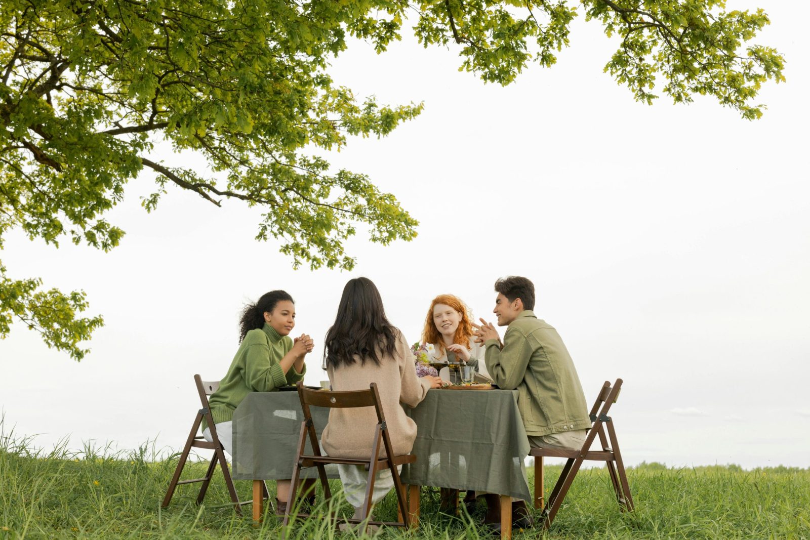 A diverse group of friends at an outdoor picnic, enjoying food and conversation under a tree.